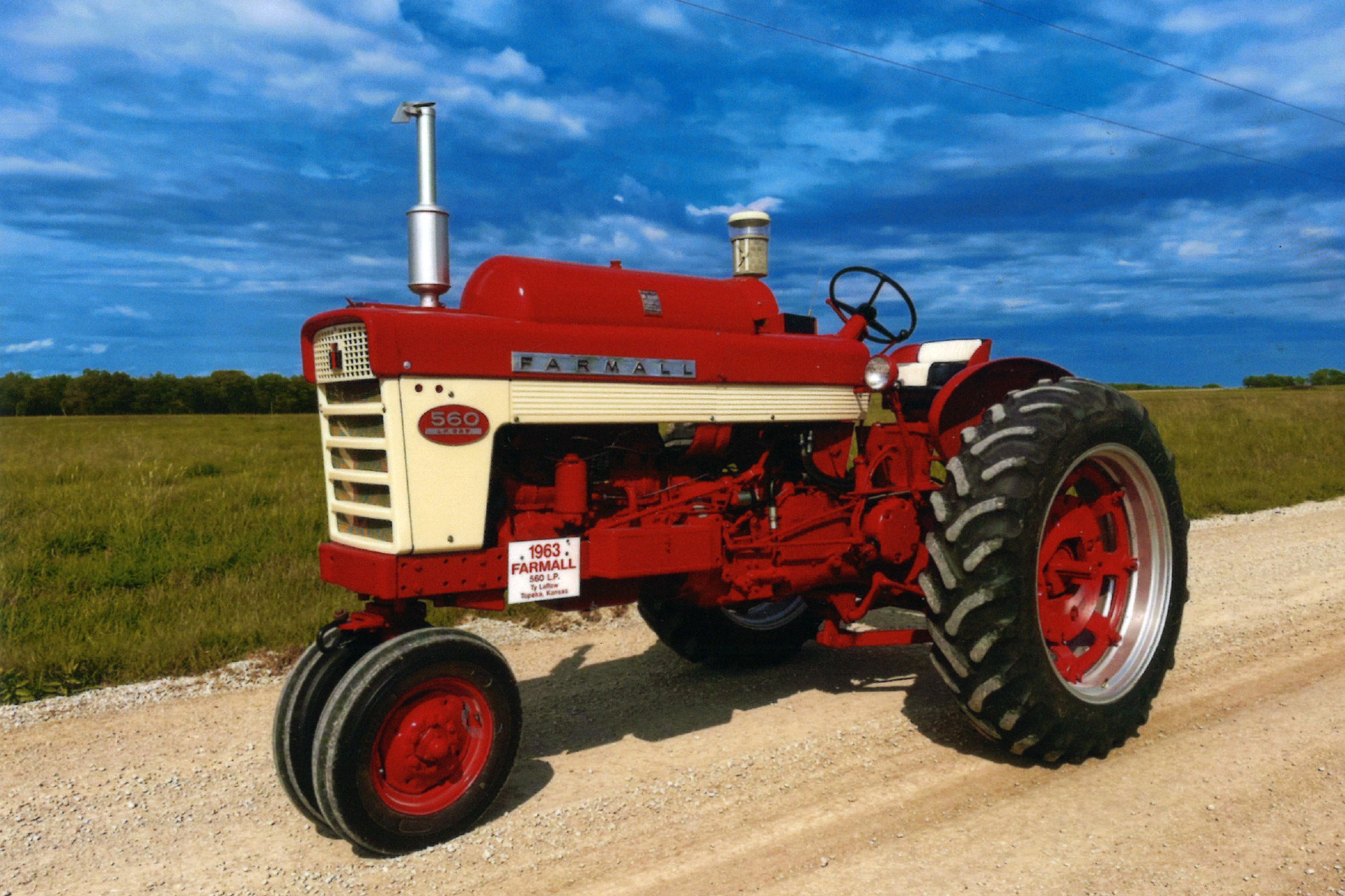 A red and white Farmall 560 tractor is parked on a gravel road with green fields and a blue sky in the background.