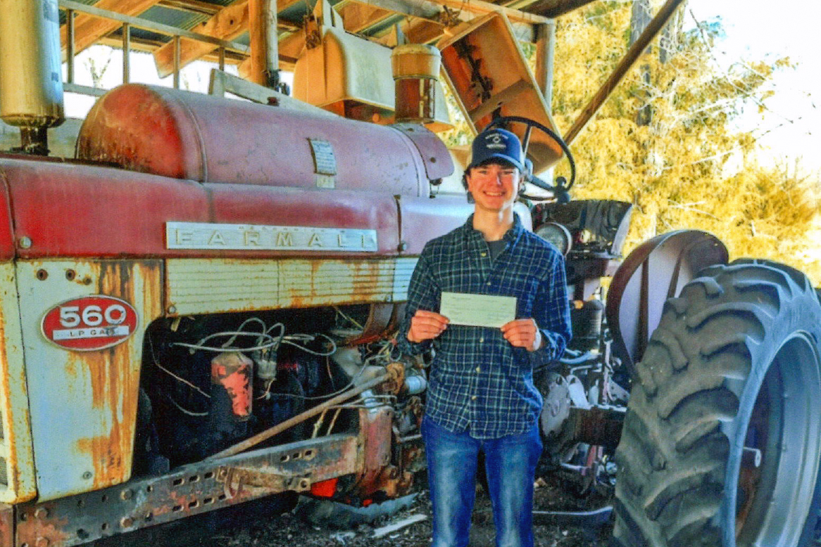 Ty La Row in a plaid shirt and cap stands smiling next to a vintage red Farmall 560 tractor inside a barn, holding a rectangular piece of paper. Sunlight and trees are visible in the background.