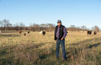 Photo of delegate Larry Triplett wearing a hat and vest stands in a grassy field with cows grazing in the background under a clear blue sky and bare trees.