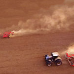 Two tractors are plowing a vast brown field. One follows the other, raising dust trails as they move across the soil. The scene is captured from above, emphasizing the scale and texture of the landscape.