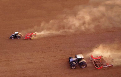 Two tractors are plowing a vast brown field. One follows the other, raising dust trails as they move across the soil. The scene is captured from above, emphasizing the scale and texture of the landscape.
