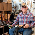 Photo of Ty May in a plaid shirt and cap is sitting in a workshop, holding a cowboy boot and tools. Several pairs of cowboy boots are displayed on the shelf behind him. The space is filled with equipment and materials for boot crafting.