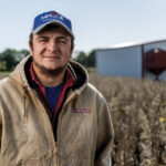 Tyler Miller wearing a blue cap and tan jacket stands in a field. He has a slight smile and a beard. In the background, there is a barn and clear sky. The field appears to be composed of dry plants or crops.