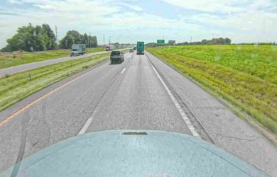 View from a vehicle driving on a highway with other cars and trucks ahead, green grassy shoulders on both sides, and a partly cloudy sky above. Signs and more vehicles are visible in the distance.