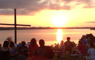 A group of people sits on benches outdoors, facing a wooden cross and a lake at sunset. The sky is partly cloudy and glows with orange and yellow hues, reflecting off the water. Trees frame the scene.