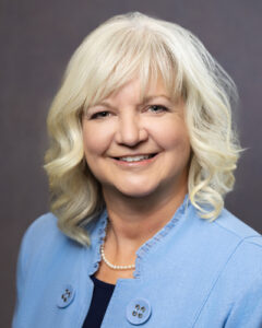 A woman with wavy blonde hair, wearing a light blue blazer, a dark top, and a pearl necklace, smiles in front of a plain, dark background.