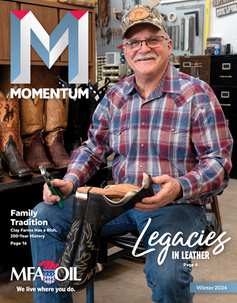 A smiling man in a plaid shirt holds a boot and a tool in a workshop filled with leather items. The magazine cover reads Momentum with features on family tradition and leather legacies.