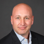 A bald man wearing a black suit jacket and a white shirt with blue stripes is smiling slightly against a dark gray background.