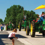 A parade of green tractors drives down a street lined with spectators. A man on the lead tractor waves while using a large yellow umbrella for shade. A young girl in a pink shirt watches from the sidewalk, holding a small flag. Trees are in the background.