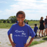 A child wearing a blue Coyote Hill t-shirt stands smiling with hands on hips in the foreground. In the background, a group of people gather on a gravel path with green fields and trees under a partly cloudy sky.