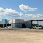 A construction site with two fuel station canopies labeled Diesel, a partially built structure, and several vehicles, including a white truck. The area is surrounded by dirt and machinery, set under a cloudy blue sky.