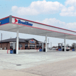 A gas station named Breaktime with multiple fueling pumps under a large canopy. The station features a convenience store in the background. The sky is partly cloudy, and the area around the station is paved and spacious.