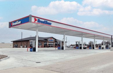 A gas station named Breaktime with multiple fueling pumps under a large canopy. The station features a convenience store in the background. The sky is partly cloudy, and the area around the station is paved and spacious.