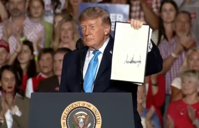 Donald Trump standing at a presidential podium, holding up a signed document to a crowd.