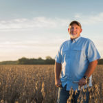 A man wearing a blue shirt and cap stands smiling in a field of crops under a clear sky. The sun is setting, casting a warm glow over the landscape.