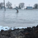 A rural scene is heavily flooded, with water nearly submerging a road sign for South 111. In the background, bare trees and farm buildings are partially visible. Sandbags and a muddy bank are in the foreground, highlighting the severe flooding.