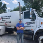 A man in a blue t-shirt and cap stands in front of a large white propane truck with American Propane Inc. and a phone number on its side. The background features trees and a partly cloudy sky.