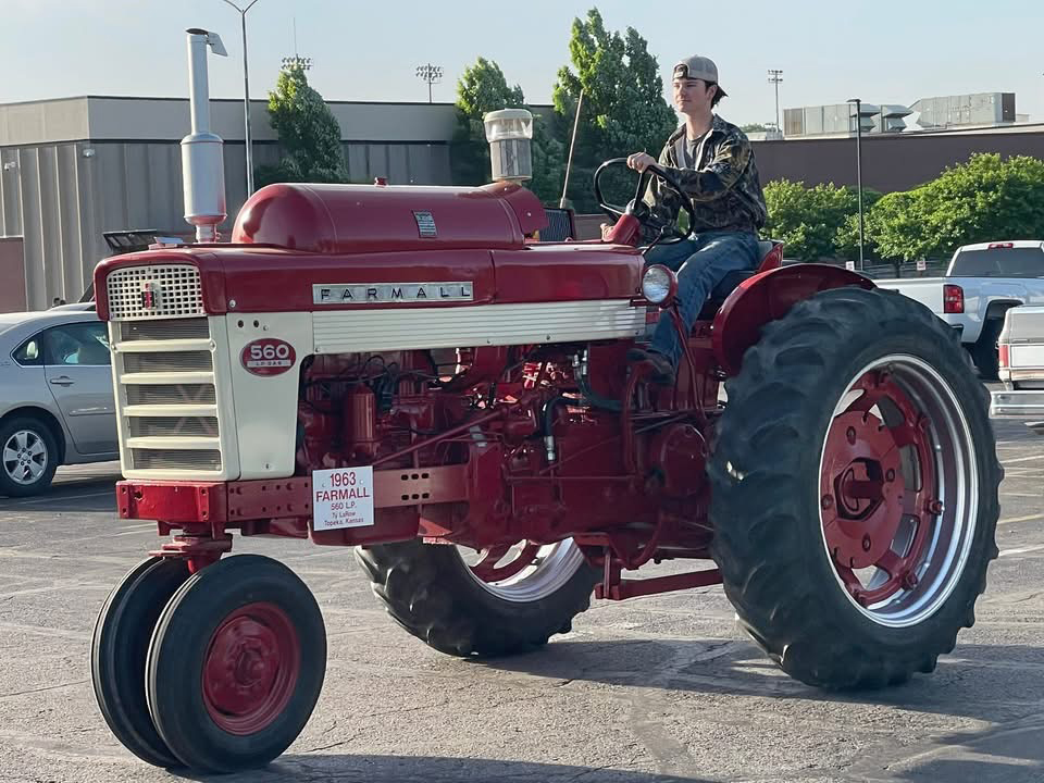 Ty La Row in a baseball cap drives a red and white 1963 Farmall 560 tractor in a parking lot with cars and buildings in the background.