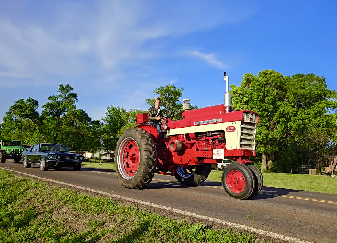 Ty La Row drives a vintage red tractor on a rural road in daylight, followed by classic cars. Trees and grass line the roadside under a clear blue sky.
