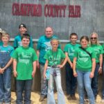 A group of children and adults in green 4-H shirts and jeans stand smiling in front of a metal wall with a red “Crawford County Fair” sign. Some wear sunglasses or hats, and one person holds a ribbon.