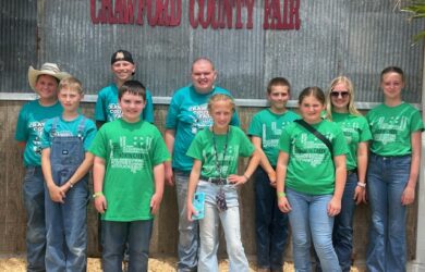 A group of children and adults in green 4-H shirts and jeans stand smiling in front of a metal wall with a red “Crawford County Fair” sign. Some wear sunglasses or hats, and one person holds a ribbon.