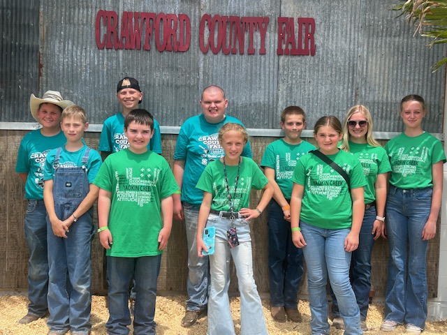 A group of children and adults in green 4-H shirts and jeans stand smiling in front of a metal wall with a red “Crawford County Fair” sign. Some wear sunglasses or hats, and one person holds a ribbon.