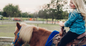 A young girl with long blonde hair, wearing a denim shirt and black pants, smiles while riding a brown pony with a blue bridle and saddle in an outdoor rural setting.