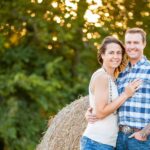 A smiling couple stands close together in front of a hay bale outdoors, with green trees and sunlight in the background. The man wears a plaid shirt and jeans; the woman wears a sleeveless top and jeans.
