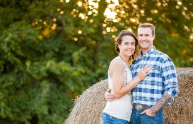 A smiling couple stands close together in front of a hay bale outdoors, with green trees and sunlight in the background. The man wears a plaid shirt and jeans; the woman wears a sleeveless top and jeans.