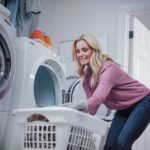 A woman in a pink sweater is mindfully loading a front-loading washing machine with her white laundry basket. Nearby, two bottles of laundry detergent rest on the dryer as she ponders over recent propane usage in the household.