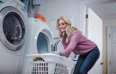 A woman in a pink sweater is mindfully loading a front-loading washing machine with her white laundry basket. Nearby, two bottles of laundry detergent rest on the dryer as she ponders over recent propane usage in the household.