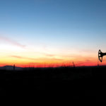 Silhouette of an oil pumpjack set against a vibrant sunset sky, with hues of orange, pink, and blue, creating a serene landscape contrast.