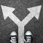 A pair of black and white sneakers stand at a fork in the road, with white arrows painted on asphalt pointing left and right, symbolizing a choice or decision-making.