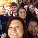 A group of young people and an adult pose for a selfie in a crowded indoor setting. Some wear navy jackets with yellow embroidered emblems. Two individuals wear paper hats with multicolored ears. They are all smiling.