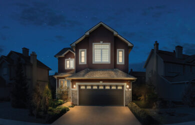 A two-story house with a brick facade and illuminated windows stands under a starry night sky. The front yard is landscaped with bushes and trees, while the driveway, flanked by exterior lights, leads to a two-car garage equipped with propane-powered generators for reliable backup power.