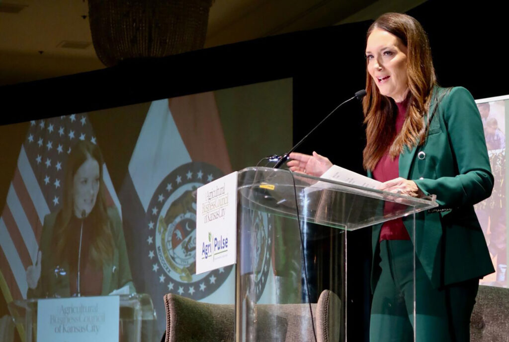 A woman in a green suit speaks at a clear podium with microphones. The podium displays signs for Agriculture Business Council of Kansas City and AgriPulse. U.S. and Missouri flags are in the background, along with a projected video of her.