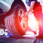 A mechanic in red overalls is adjusting a car tire on a hydraulic lift in a brightly lit garage. The sun streams in from the background, illuminating the scene. A toolbox is positioned nearby in the workshop.