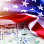 A close-up of the American flag draped over a pile of U.S. dollar bills, featuring various denominations. The flags stars and stripes are clearly visible, contrasted against the currency below. Light shines softly over the scene.