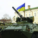An armored military vehicle with a mounted weapon displays the Ukrainian flag. It is parked in front of a beige, multi-story building with a green roof. The background includes bare trees under a cloudy sky.