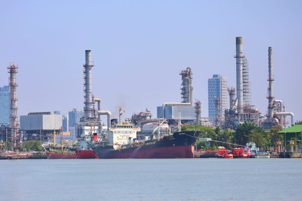 Large cargo ships docked at an industrial port with oil refinery towers and structures in the background, under a clear blue sky.