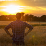 A person stands in a field with tall grass, facing a vibrant sunset. They wear a plaid shirt and have their hands on their hips. The horizon shows silhouetted trees under a glowing sky with shades of orange and yellow.