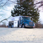 A blue and silver oil delivery truck is parked on a snowy road. Trees with bare branches frame the scene under a clear sky. The truck has MFA Oil written on the side.