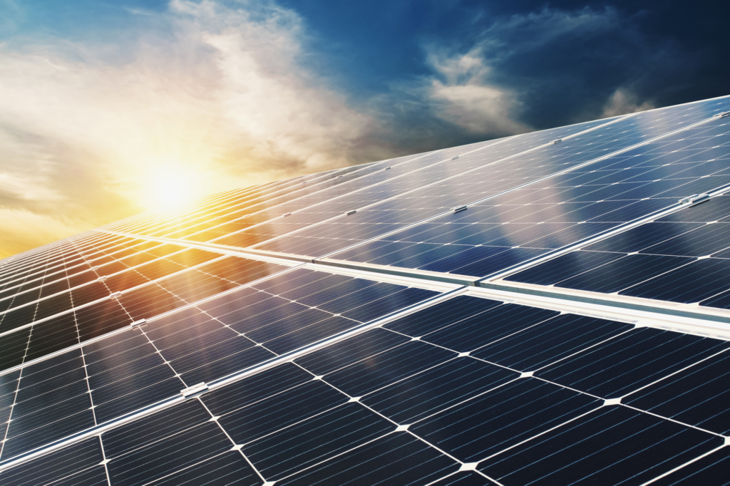 A close-up of a solar panel array reflecting sunlight. The sun is partially behind clouds, creating a dramatic sky with bright and dark areas. The panels are positioned at an angle, capturing the suns rays.