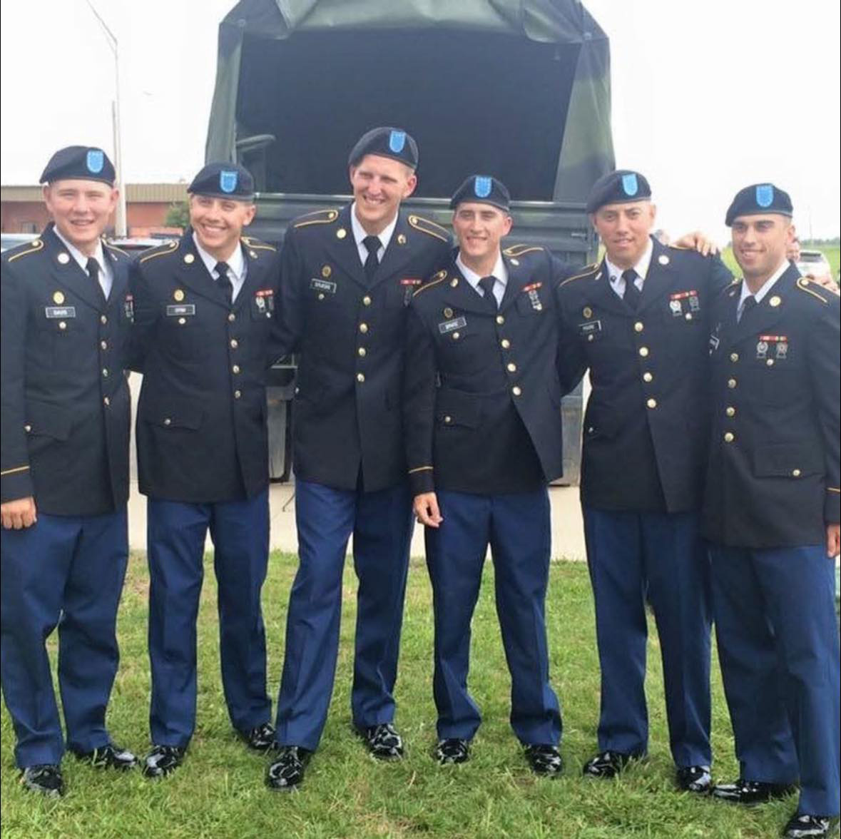 Six men wearing U.S. Army dress uniforms and berets stand in a row outdoors, smiling in front of a military vehicle. They have their arms around each other and appear to be celebrating or posing for a group photo.