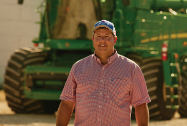 A person in a pink checkered shirt and blue cap stands in front of a large green agricultural machine on a sunny day. The background includes a metal silo.