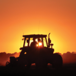 Silhouette of a tractor in a field during sunset, with a vibrant orange sky and sun setting behind it. Dust trails follow the tractor, and faint outlines of trees and a wind turbine are visible on the horizon.