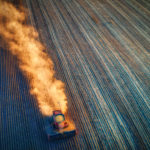 Aerial view of a red tractor plowing a vast field, leaving a trail of dust behind. The field is lined with parallel furrows, creating a striped pattern. The lighting emphasizes the texture and colors of the soil.