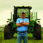 A man in a cap and blue shirt stands with arms crossed in front of a large green agricultural machine on a grassy field. Overcast sky in the background.