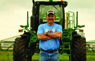 A man in a cap and blue shirt stands with arms crossed in front of a large green agricultural machine on a grassy field. Overcast sky in the background.